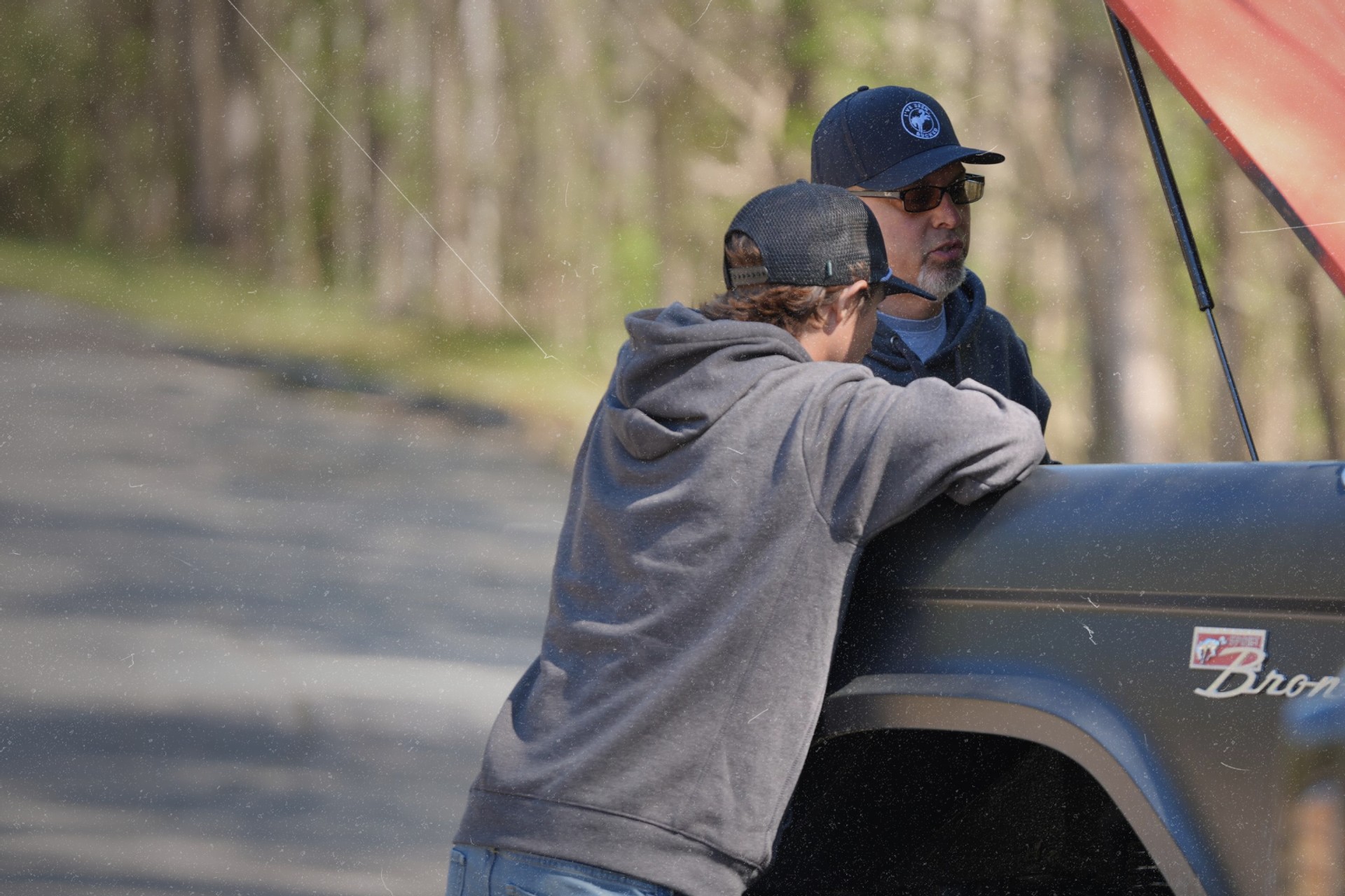 Man in IBB trucker hat drinking from a mug by a campfire, Bronco in background